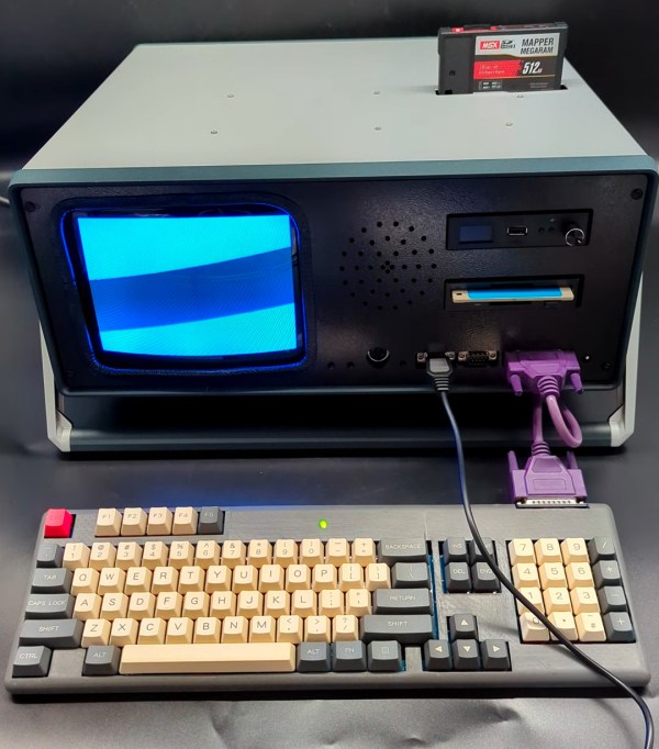 A "portable" computer in a grey enclosure. There is a small CRT on the left hand side of the face of the enclosure and a disk drive and a couple ports exposed on the right hand side. The keyboard is attached with a purple cable. A black cartridge with a grey and red label sticks out of the top of the enclosure.