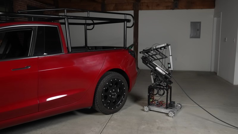A red Tesla Model 3 converted into a pickup truck with a black lumber rack extending over the roof of the cab sits in a grey garage. A black and silver charging robot is approaching its charging port from the right side attached to a black cable. The charging bot is mostly a series of tubes attached to a wheeled platform and the charging connector itself is attached to a linear actuator to insert the charging device.