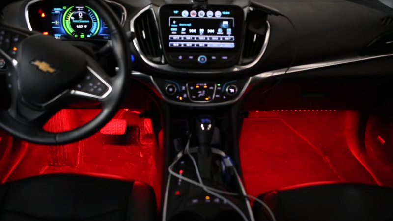 A view of the inside of a car, with drivers wheel on the left and control panel in the middle, with red LED light displayed in the floor area under the drivers wheel and passenger side.