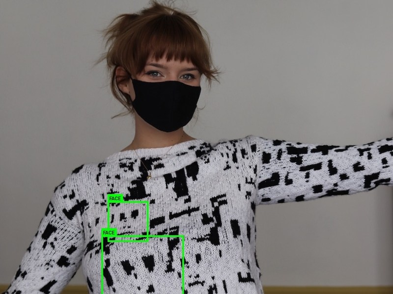 A woman with red hair in a black and white jumper/sweater. Two green blocks outline sections of the pattern saying "face" as indicated by a face detection system. The woman's face is covered by a mask and not detected.