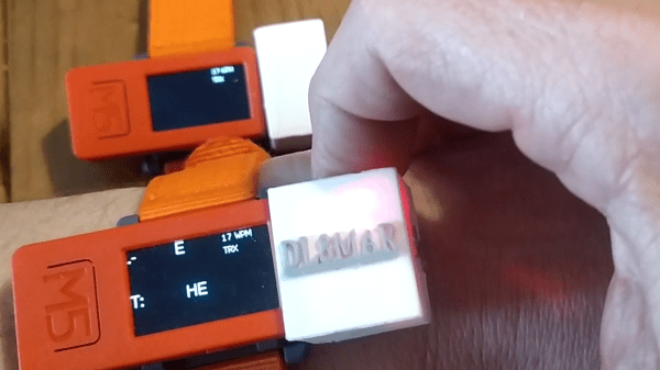 An orange m5stickc plus strapped to a wrist in the foreground with the persons other hand pressing down on the top of an installed hat, communicating with another m5stickc plus in the background on the table.