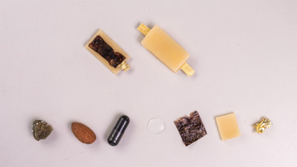 A series of food items along the bottom of the frame including an unidentified grey block, an almond, a food supplement capsule, a square of seaweed, a square of beeswax, and a crumpled up piece of gold foil. At the top of the image is a fully assembled battery with electrodes sticking out the ends of a block of beeswax and a half finished battery with the nori separator visible.