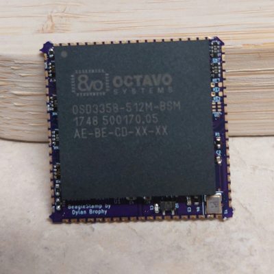 A populated purple PCB propped against a piece of wood on a light grey work surface. The bulk of the PCB is covered in an Ocatavo processor chip.