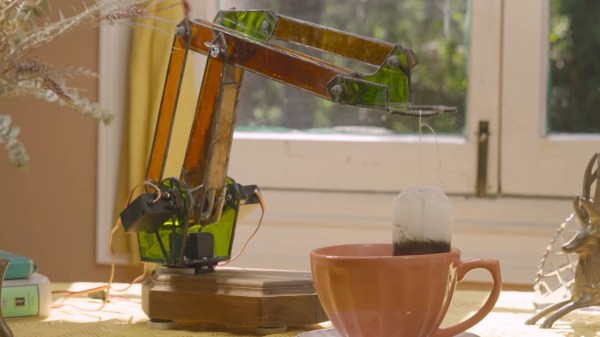 An orange and green stained glass robot arm sits on a table with a yellow lace tablecloth. It suspends a teabag over a brown teacup. You can see green leaves outside the window behind the bot.