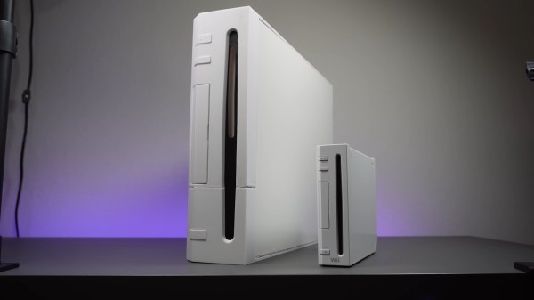 A white Wii console sits on a grey table in the vertical orientation with its front facing the camera and its back away from the camera at a slight angle to the right. Next to it is a 2x sized replica which dwarfs the diminutive console. A purple light runs across the back edge of the table.