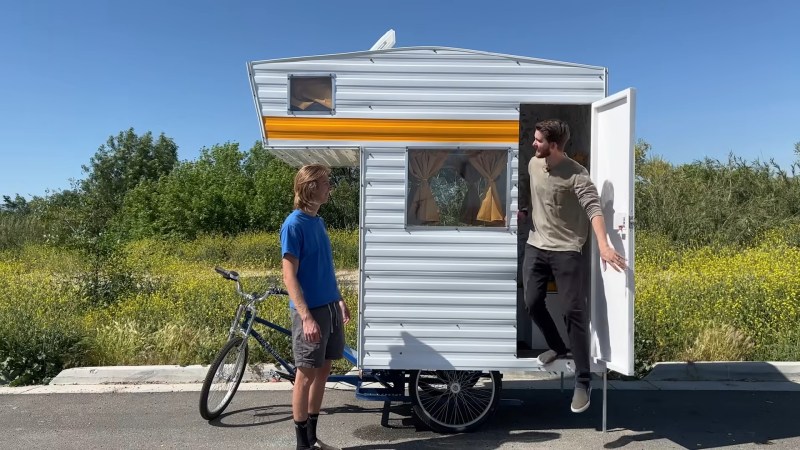 A blue work tricycle (single front wheel with platform between two rear wheels) sits on an asphalt path with trees and wildflowers in the background. A boxy, white camper with a front overhanging the bike seat is attached to the platform. It has a yellow stripe and curtains like many campers did in the 1970s. One white male is exiting the door of the camper while his brother is standing to the left of the camper by the bike seat.