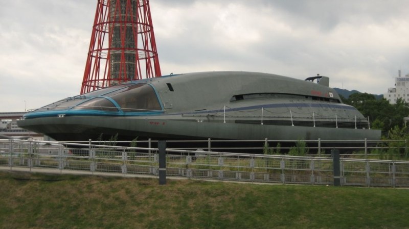 The Yamato 1, a sleek grey ship that looks vaguely like a computer mouse or Star Trek shuttlecraft. It has an enclosed cockpit up front with black windows and blue trim. It is sitting on land in front of a red tower at a museum in Tokyo.