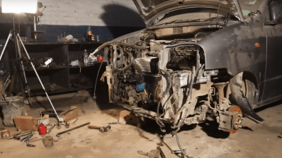 An open engine bay for a small car. The wheels are off so the hubs and brakes are visible to the side and the electric motor mounted on top of the vehicle's original engine block is in the center of the image.