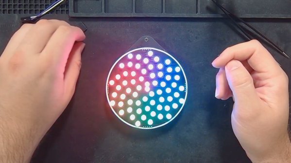 A black work mat holds a circular badge with 64 addressable LEDs in a spiraling shape akin to the center of a sunflower. The LEDs have a rotating rainbow spiraling around the circle with red touching violet on one end. The colors extend in bands from the center to the rim of the circle.