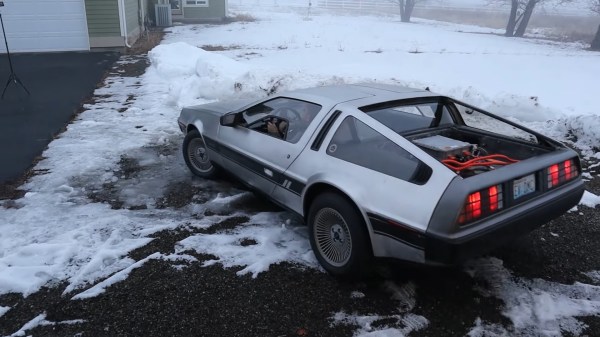 A DeLorean sitting on patchy snow next to a driveway. It's angled away from the viewer to the left showing off the open engine compartment with bright orange high voltage lines coming out of a square metallic charger box.