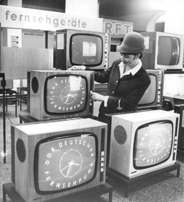Black and white photograph of a display of televisions displaying a DDR Deutsche Frensehfunk logo, with an attendant adjusting one of the sets.