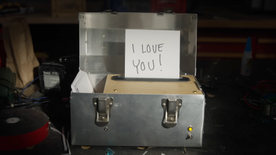 An aluminum box sits on a workbench. It is open and has a message saying "I Love You!" inserted in a wooden slot. There is a switch with a yellow LED on the front and a small compartment to the left of the wooden slot to store paper.