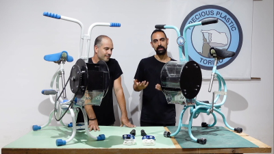 Two men in black shirts stand between a white and a blue exercise bike sitting on a table in front of them. The exercise bikes have black drums slightly larger than a coffee can affixed to the front of the bike which houses the shredding mechanism. In the background is a "Precious Plastics Torino" circular logo.