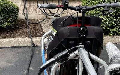 A silver front loader cargo bike sits in a parking lot in front of an electric vehicle charger. A cable runs from the charger to the bike.