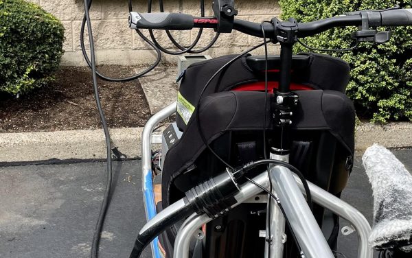 A silver front loader cargo bike sits in a parking lot in front of an electric vehicle charger. A cable runs from the charger to the bike.