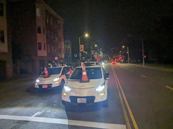 Two white Chevy Bolt hatchbacks sit side-by-side, immobilized in the street, their roofs festooned with sensors and an orange cone on their hoods like a snowman's nose pointed toward the sky.