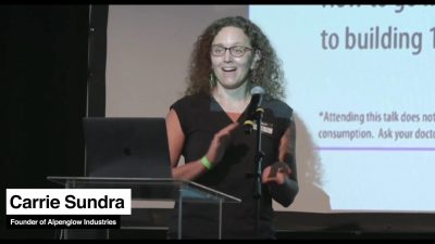 A woman with a black vest and pink shirt with curly hair stands behind a podium in front of a projected presentation. She is speaking and has her hands moving in a vague guesture.