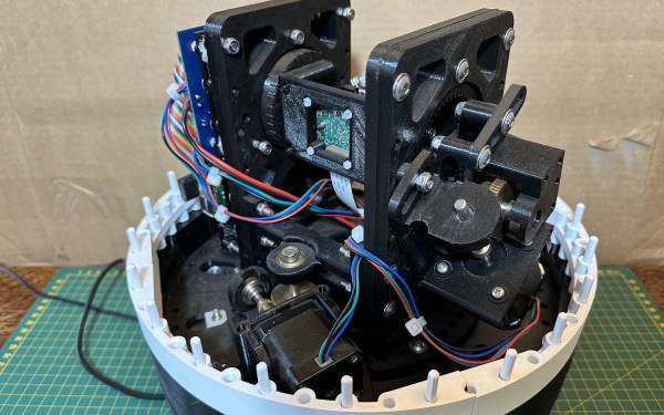 A black motion system with two stepper motors. A green circuit board is fixed in a rotating cage in the center, and the entire assembly is on a white base atop a green cutting mat. Wires wind through the assembly.