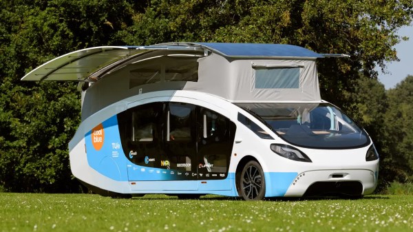 A large, teardrop-shaped van with a wide, friendly face sits in a grassy field. A grey canvas pop top is opened on its top and solar panels extend from either side of its roof, making it look somewhat like a large insect with wings extended.