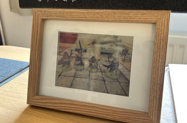 A brown, wooden picture frame with a white matte holds a slightly pixelated photo of gaming miniatures. It is sitting on a wooden table.