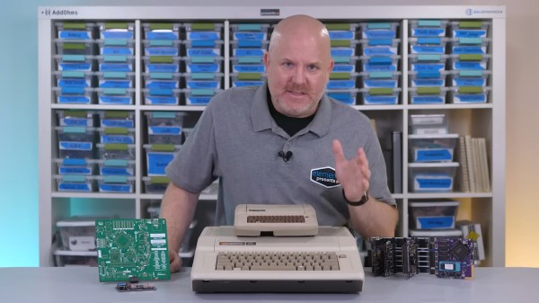 A bald white man stands behind a table with an Apple II, a large green PCB, and a modular purple and black development board system. Atop the Apple II is what appears to be a smaller Apple II complete with beige case and brown fake keyboard.