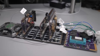 A set of PCBs sits on a table. There is a logic analyzer plugged into one end that looks like a grey square. Three boards stick up at right angles from the main plane which consists of a purple square PCB with the IIe ROM and MEGA chips and a black rectangular PCB with four sets of headers for PCB modules to slot into.