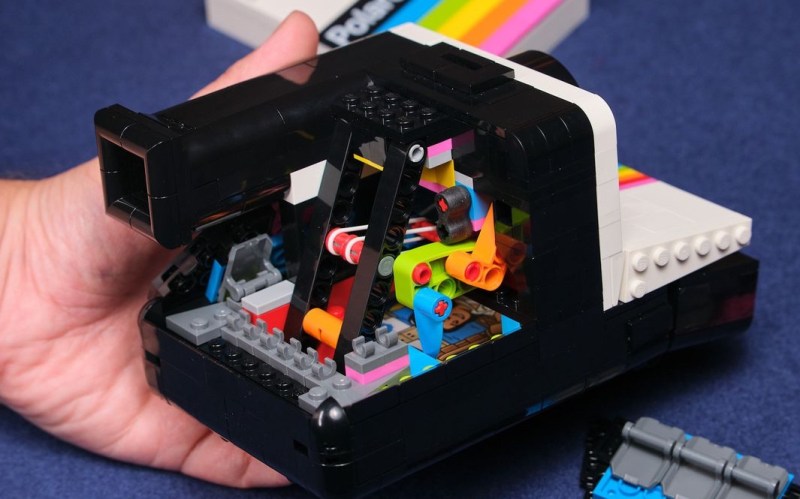 A hand holds a LEGO replica of a Polaroid camera. The back of the "camera" has been removed to show the sereies of Technic pieces inside that allow the camera shutter to work.