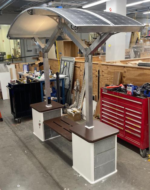 A dark brown bench suspended between two white and grey rectangular pillars. They are capped in the same brown HDPE material. Aluminum uprights go to a curved solar panel roof that looks somewhat similar to a paragliding chute. The bench is inside a clean-looking workshop with two large toolboxes against a plywood half wall.