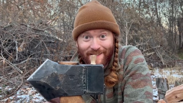 A white man with red hair in pigtails under a brown cap holds an axe with a black head and wooden handle. The axe has a rectangular box welded onto the back side of its trapezoidal head.