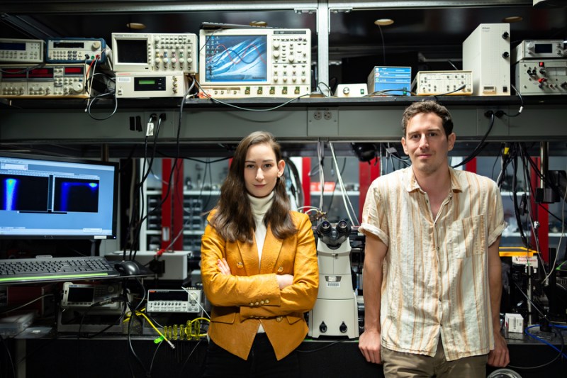 Team members Madeleine Laitz, left, and lead author Dane deQuilettes stand in front of a tidy lab bench equipped with oscilloscopes and computers. Laitz has a snazzy yellow jacket that pops compared to the neutrals and blues of the rest of the picture.