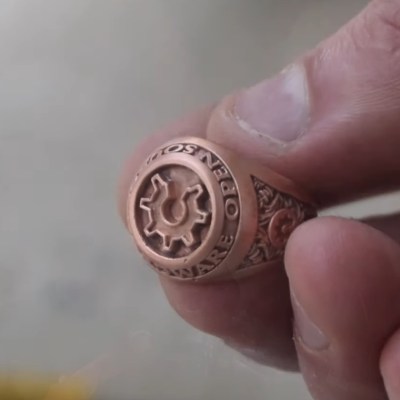 A hand holds a very detailed copper ring. It is inscribed with the words "Open Source Hardware" and the open gear logo associated with open source hardware. It looks kinda like a class ring.
