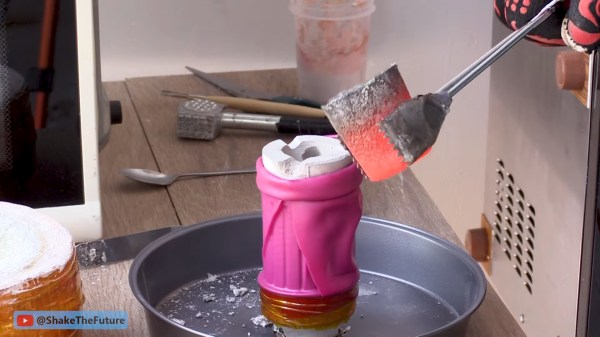 A red hot crucible is held with metal tongs above a white plaster mold. The mold is held in a bright pink silicone sleve atop a metal pan on a wooden workbench. Red cheese wax holds the sleeve to a metal funnel connected to a vacuum cleaner.