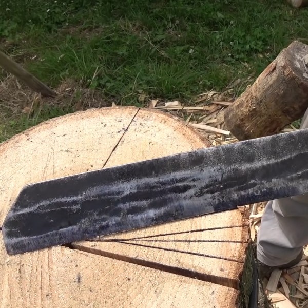 A man standing next to a log holds a wooden mallet and a grey froe with a wooden handle. The froe's long straight blade sits atop the end of the log. Several cuts radiate out from the center of the log going through the length of the wood.