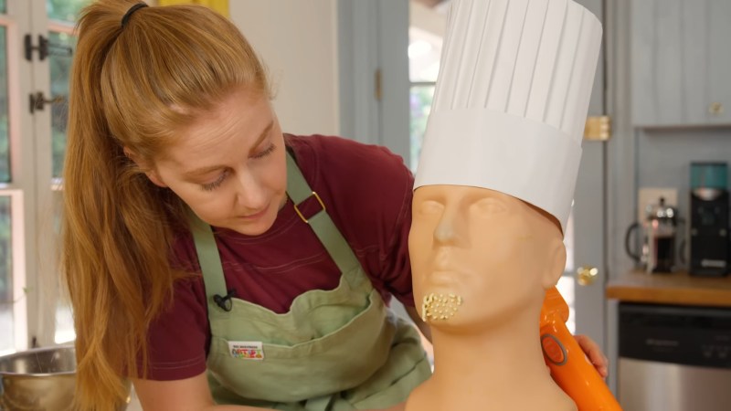 A white woman with a long ponytail in a green apron looks down at a mannequin head with pasta coming out of its chin. There is an orange pasta gun sticking out of the back of its head and a chef's hat on its head. It looks vaguely like a bust of Ramses.