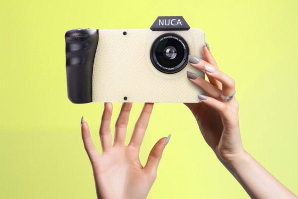 A pair of hands holds a digital camera. "NUCA" is written in the hood above the lens and a black grip is on the right hand side of the device (left side of image). The camera body is off-white 3D printed plastic. The background is a pastel yellow.
