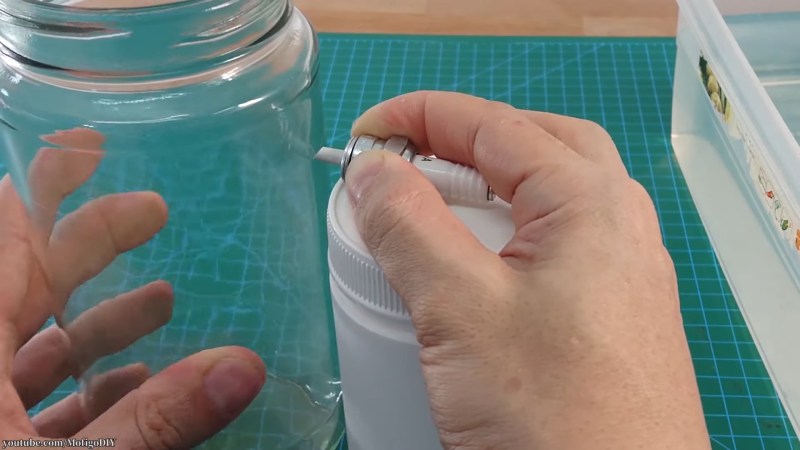 A person holds a glass jar in their left hand and a spark plug in their right atop a white cylindrical canister. The jar and canister are sitting on top of a green cutting mat.