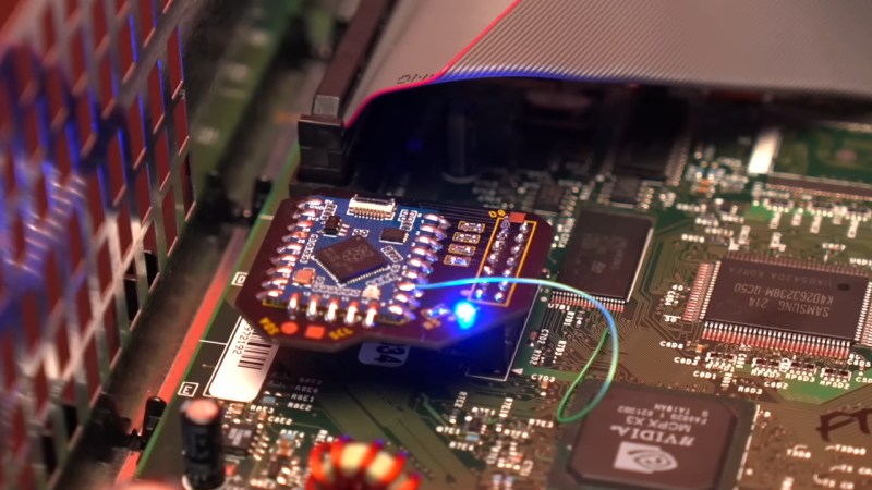 Showing the modchip installed into a powered up Xbox, most of the board space taken up by a small Pi Pico board. A wire taps into the motherboard, and a blue LED on the modchip is lit up.