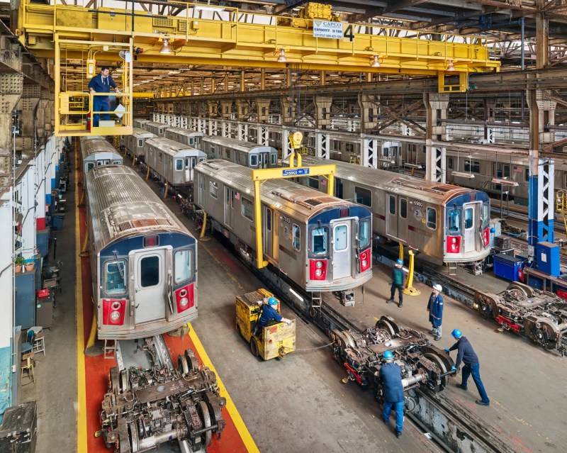 A warehouse with concrete floors and at least four subway car rails running off into the distance. On the rails are dozens of R142 series subway cars with refurbished trucks in the foreground. People are visible on the floor moving a truck, and one man is in a bright yellow crane above everything watching what happens.