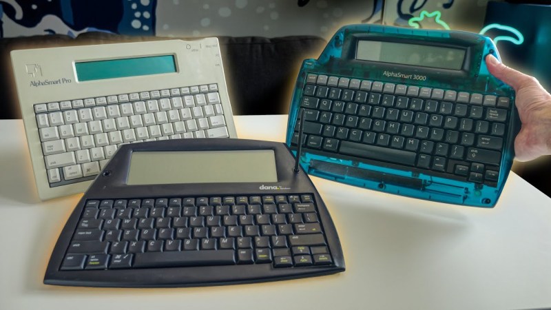 Three different keyboard devices with greyscale LCDs on top are on a white table. In the back left is a beige one with grey keys, front and center is a black model, and to the right is a translucent blue model with black keys being held up by a hand.