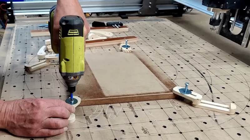 A person putting a screw into a CNC spoil board on the left of the image. Their drill is chartreuse and black. Clamps hold a rectangular board down at all four corners. The spindle of the CNC is just visible on the right hand side of the image.