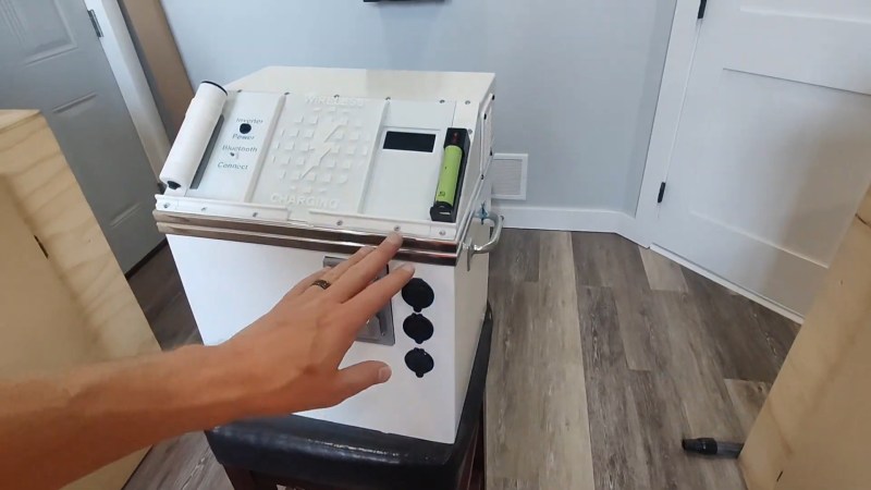 A man's hand with a black ring touches a white box. It is square on the bottom and has a sloped top. Various AC, 12V, and USB ports adorn its surface. It's approximately the size of a human head.