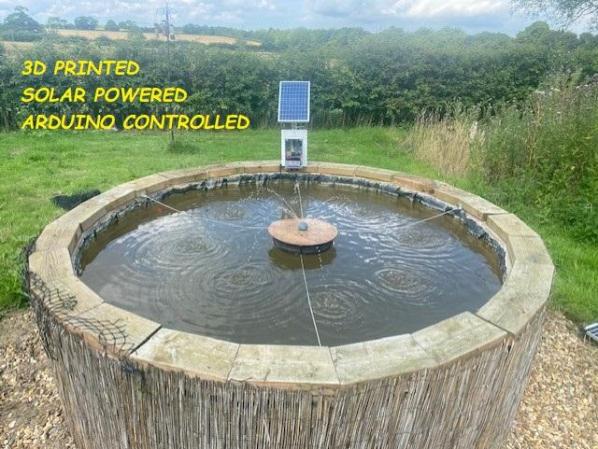 A circular concrete pond in a garden. A small round fountain jets water out in the center and a solar panel and control box are visible attached to the end of the pond opposite the camera. On the top left is the text, "3D printed, Solar powered, and Arduino controlled" in yellow