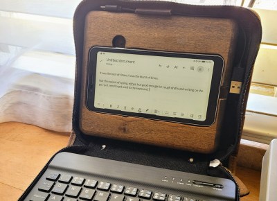 A black and brown planner opened like a laptop. Inside is a greyscale eink phone inside a brown piece of MDF on top, and a black keyboard fills the bottom half.