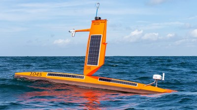 A bright orange sailboat with solar panels on the wing sail and the hull of the craft. A number of protuberances from the wing are visible containing instruments and radio equipment.