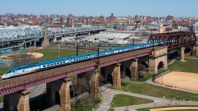 A new blue and white high speed train crosses a brick bridge. There is what looks like a park beneath and a cityscape in the background.