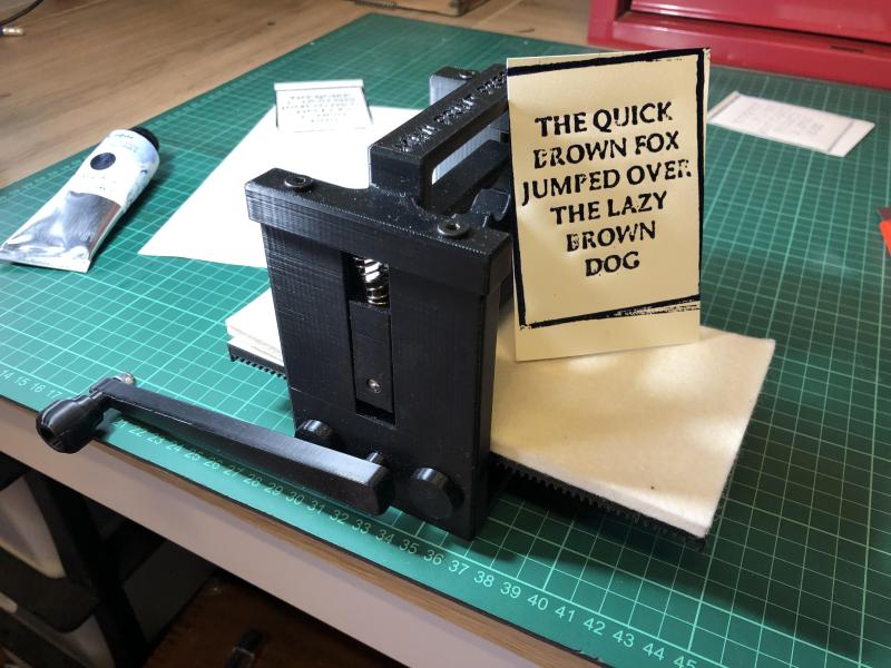 A small 3D-printed printing press with a print that says THE QUICK BROWN FOX JUMPED OVER THE LAZY BROWN DOG.