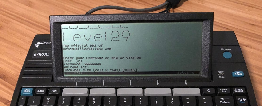 A black device with a monochrome LCD sits on a wooden table. It's keyboard extends below the frame. On the screen is the "Level 29" BBS service login.