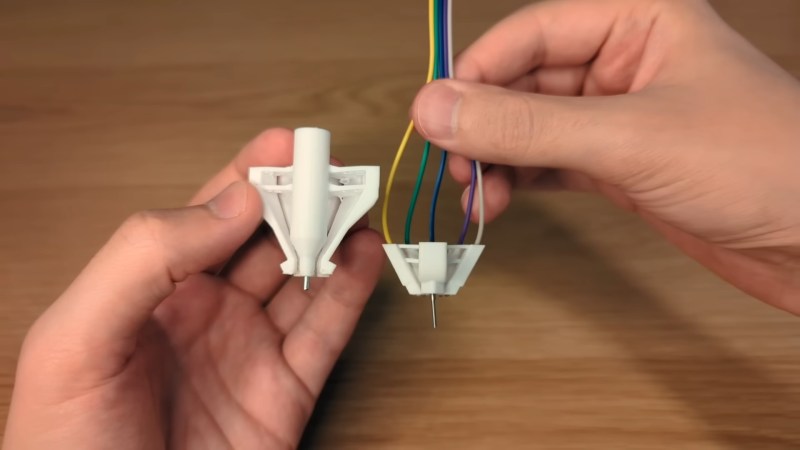 Hands hold a set of white, 3D printed connectors above a wooden table. They look like a cross between a ballpoint pen tip and a spider. The shorter one on the right has yellow, green, black, purple, and white wires coming out the top.