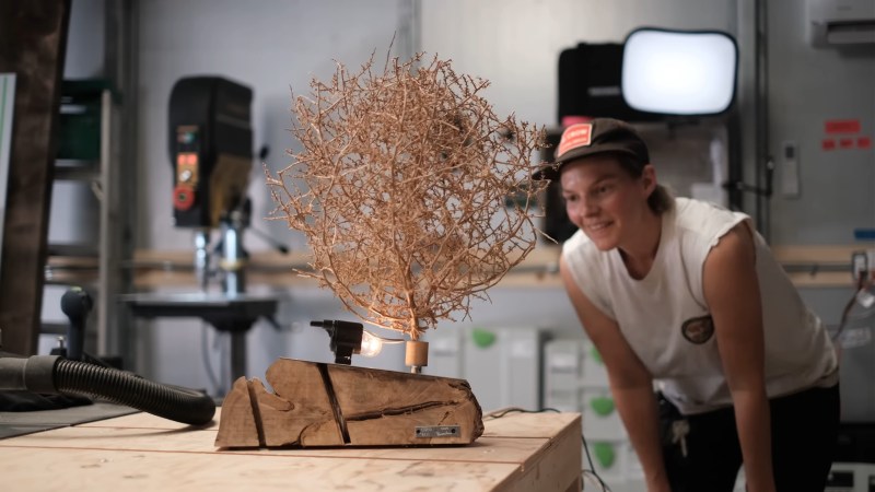 A rough cut piece of wood sits on a workbench. A light and a tumbleweed are mounted on top so that the light shines through the tumbleweed. A woman in a ball cap and white tank top is crouched in the background smiling.