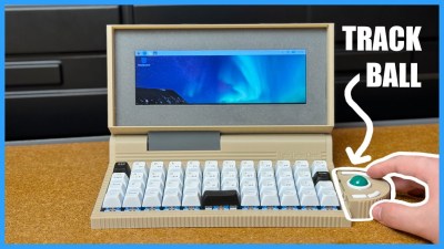 A very wide beige laptop sits on a wooden table. A hand manipulates a teal ball in a semicircle attached to the right sided of the device. The track ball and hand are outlined in white.